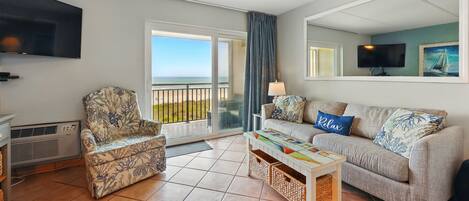 Una sala de estar con un sofá, un sillón y una mesa de centro. Hay un televisor montado en la pared y una ventana con vista a la playa.