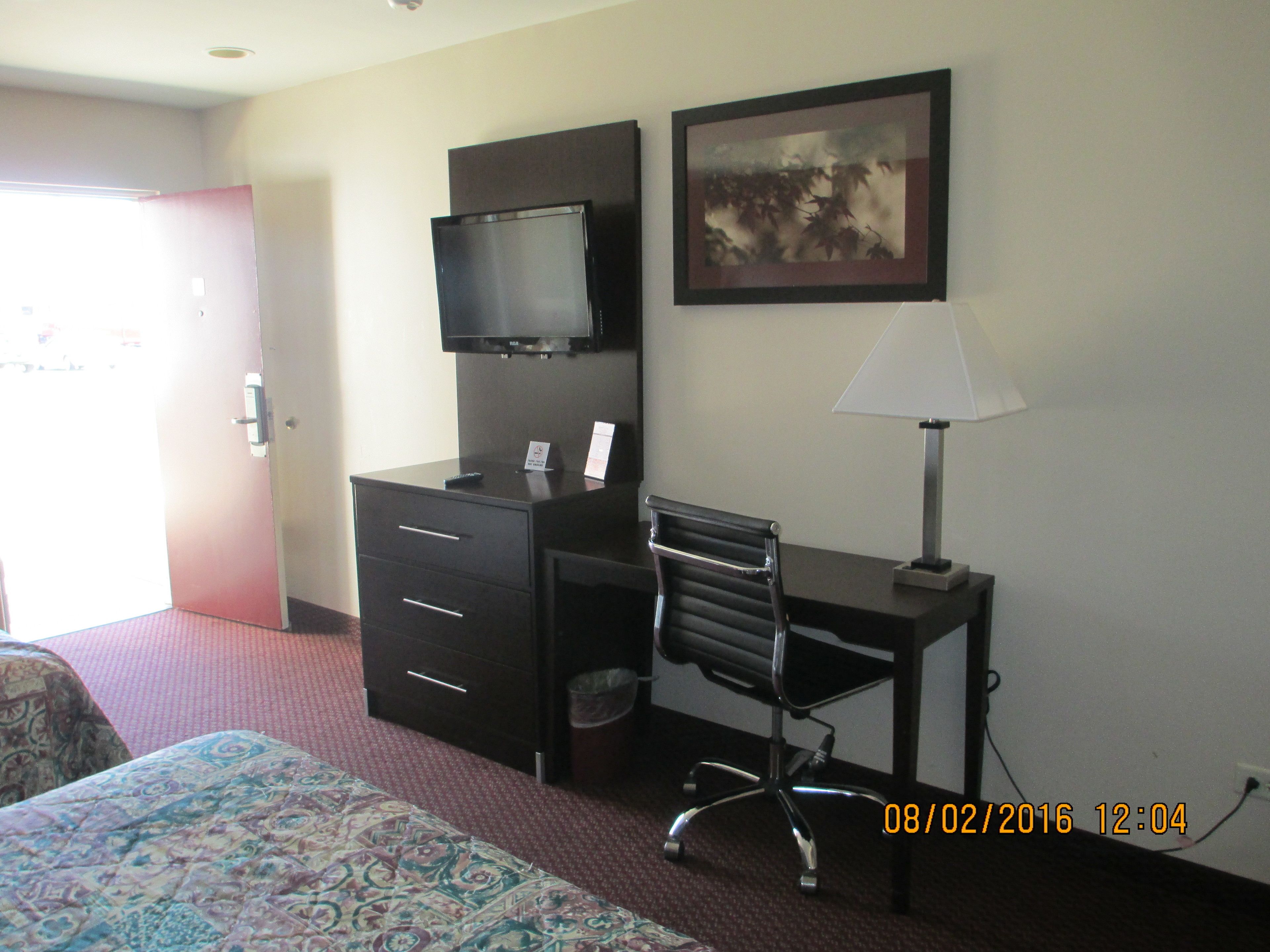 A hotel room with a bed, a desk with a chair, a television, a lamp, and a painting on the wall.