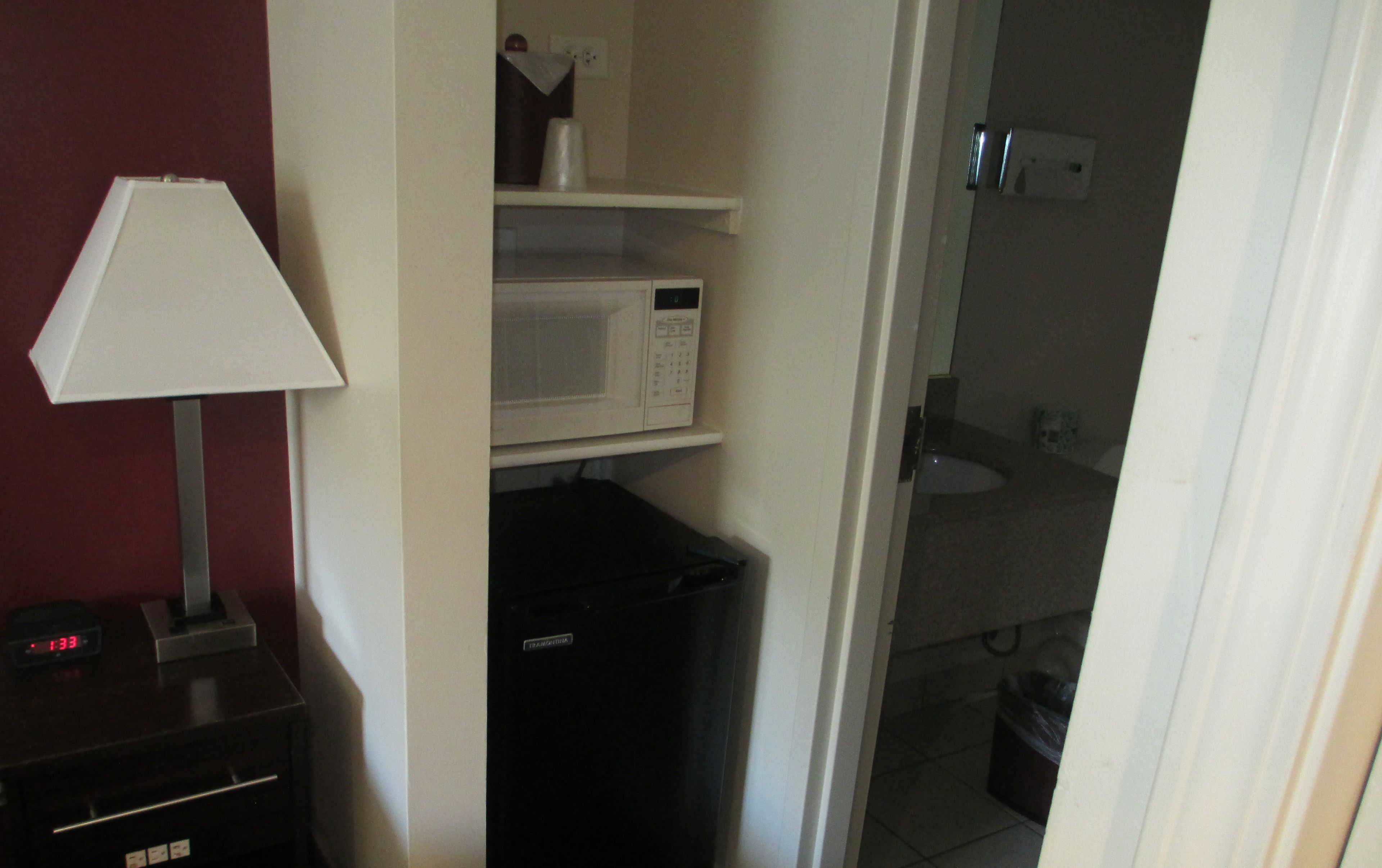 Habitación compacta con microondas en un estante, un refrigerador pequeño, una lámpara y un baño visible a través de una puerta abierta.