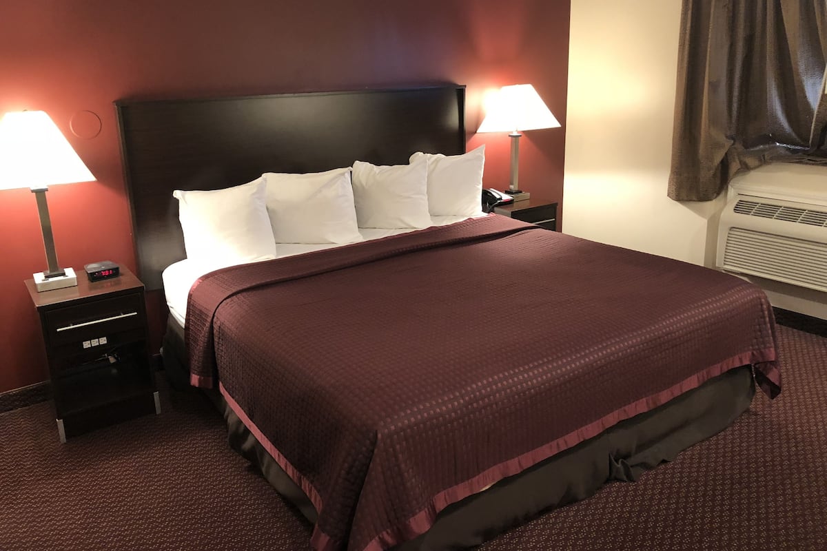 A hotel room with a large bed, two bedside lamps, a nightstand, a thermostat, and a window with curtains.