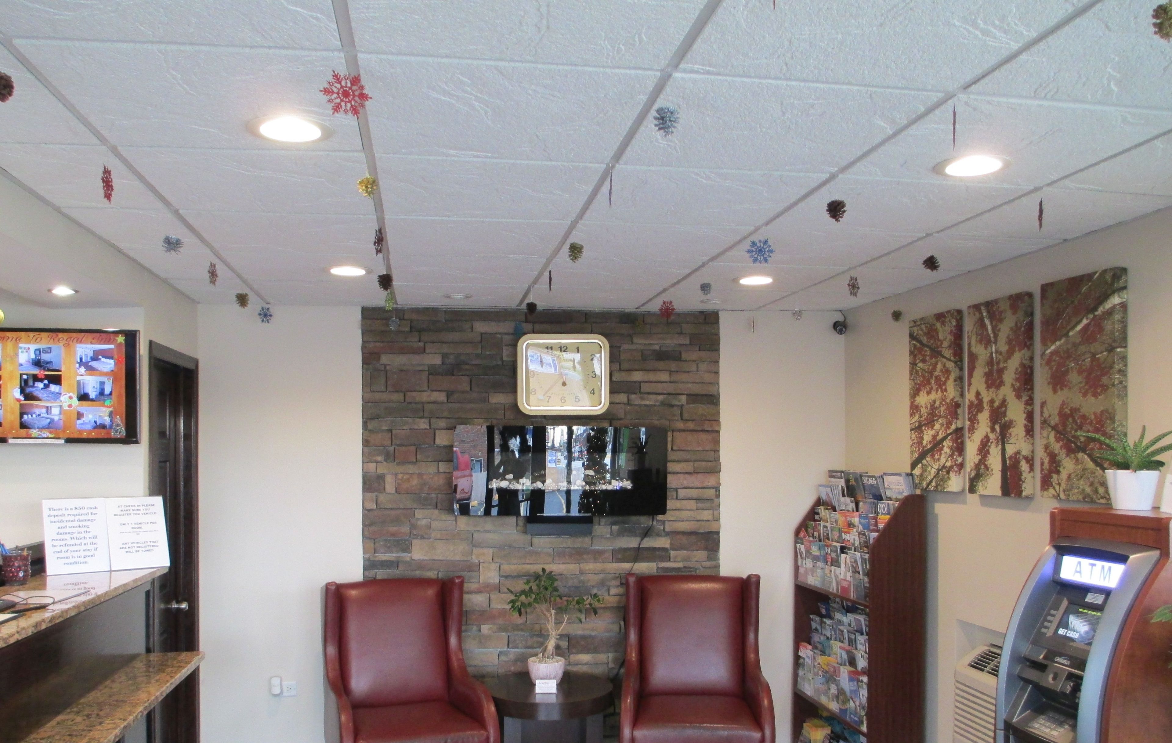 A lobby with a brick wall, a clock, a TV, two leather chairs, a small table with a plant, and an ATM.
