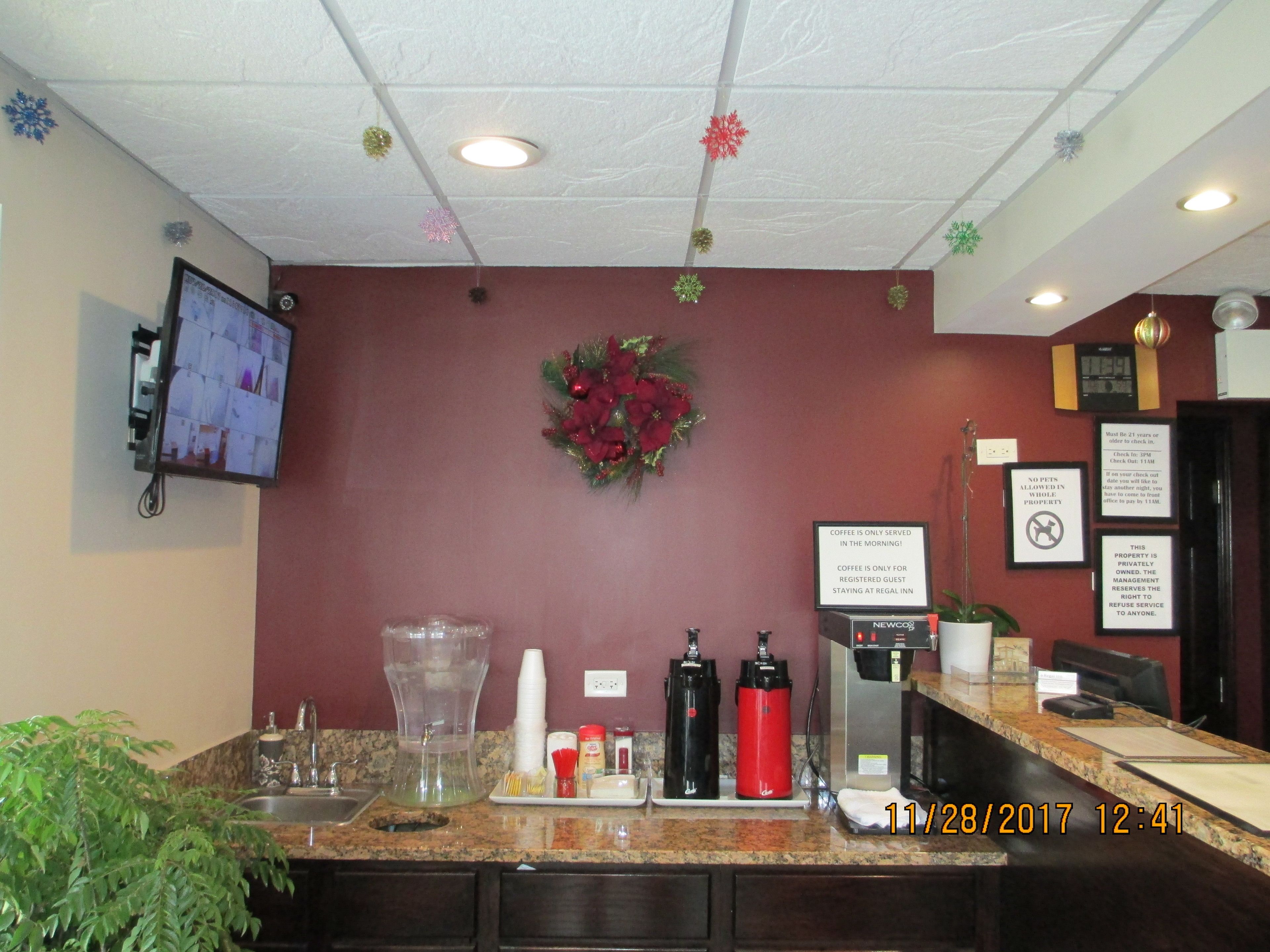 A hotel reception area with a coffee station, a monitor displaying a room, and a wall with holiday decorations.