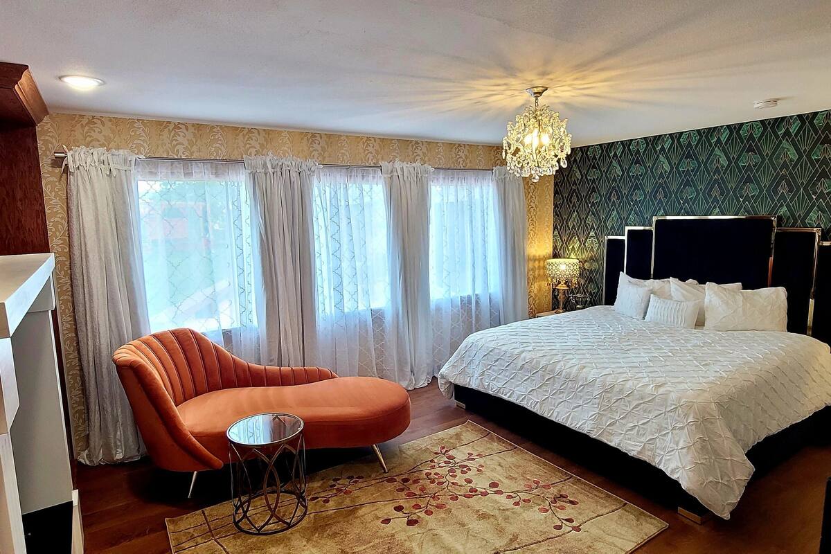 A bedroom with a bed, a chair, a small table, a chandelier, and a patterned wall.