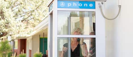A public phone booth with a clear glass window, situated in front of a building with a red-tiled roof and a green door.