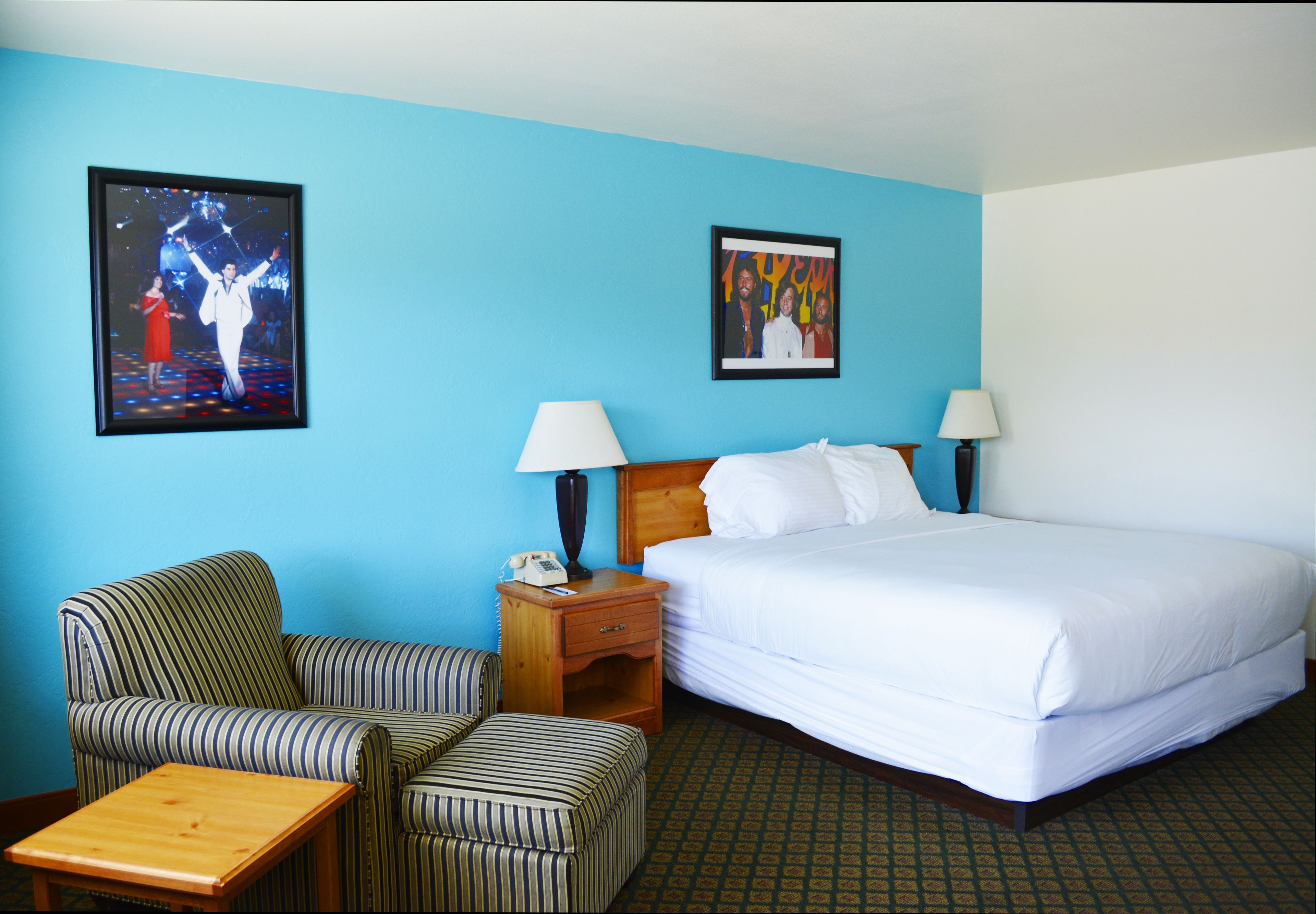 A hotel room with a bed, two bedside tables with lamps, a striped armchair, a small wooden table, and two framed pictures on the wall.