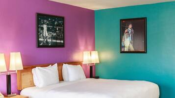 Habitación de hotel con una cama grande, dos mesitas de noche con lámparas, un tocador de madera, un banco y dos cuadros enmarcados en las paredes.