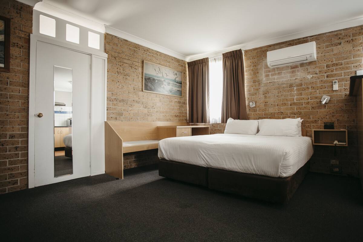 A hotel room with a brick wall, a bed with white linens, a wooden desk, and a window with curtains.