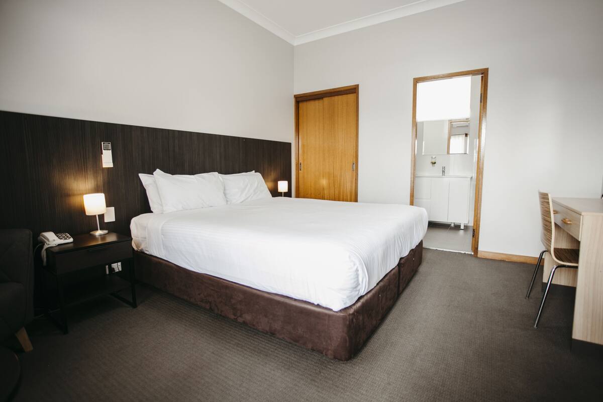 A hotel room with a large bed, a wooden headboard, a desk with a chair, a nightstand with a lamp, and a wardrobe.