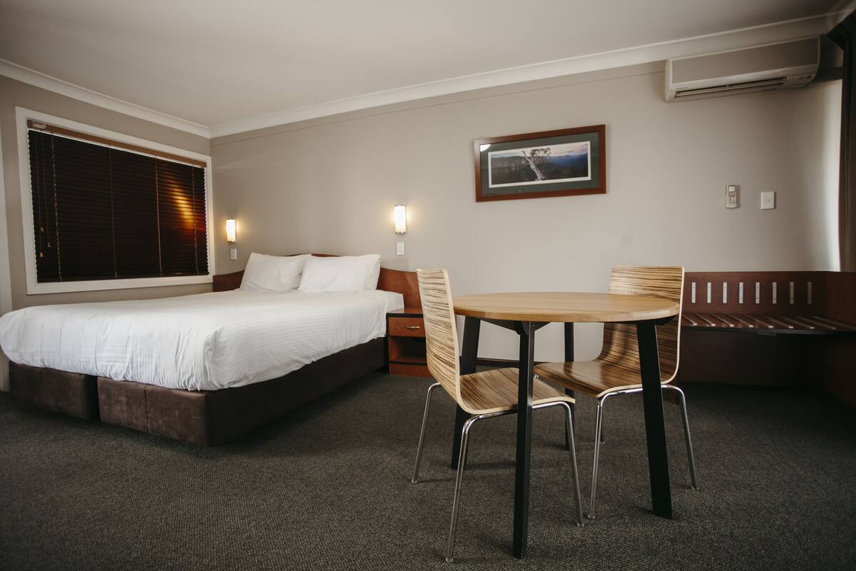 A hotel room with a bed, a table with chairs, a bench, and a wall-mounted picture.