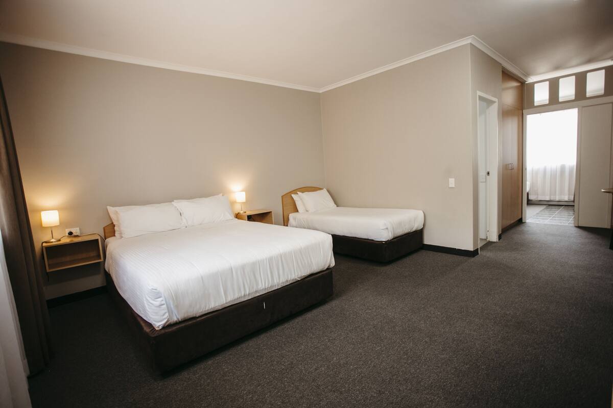 A hotel room with two single beds, a nightstand with a lamp, and a door leading to another room.
