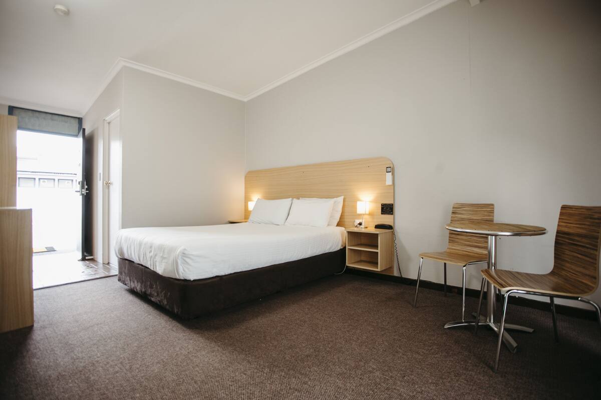 A hotel room with a bed, a nightstand with a lamp, a wooden chair, and a small table.