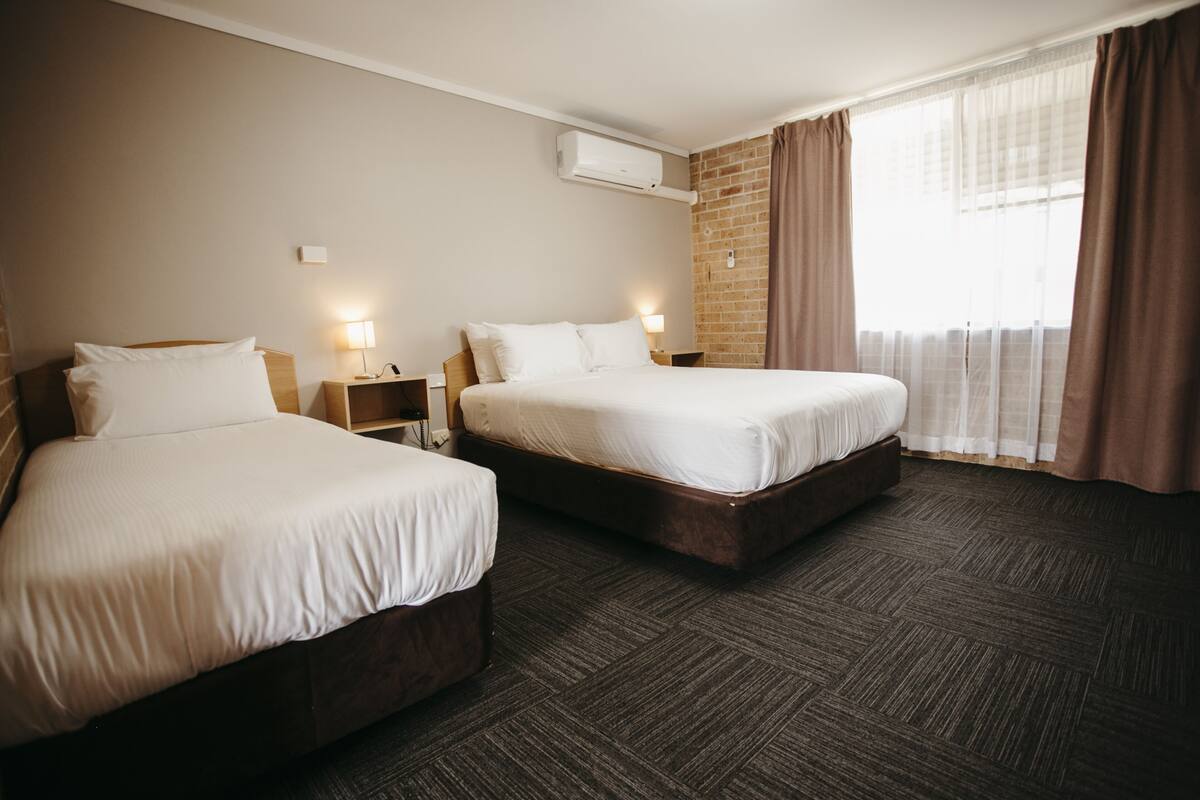 A hotel room with two beds, a brick wall, an air conditioning unit, and a window with curtains.