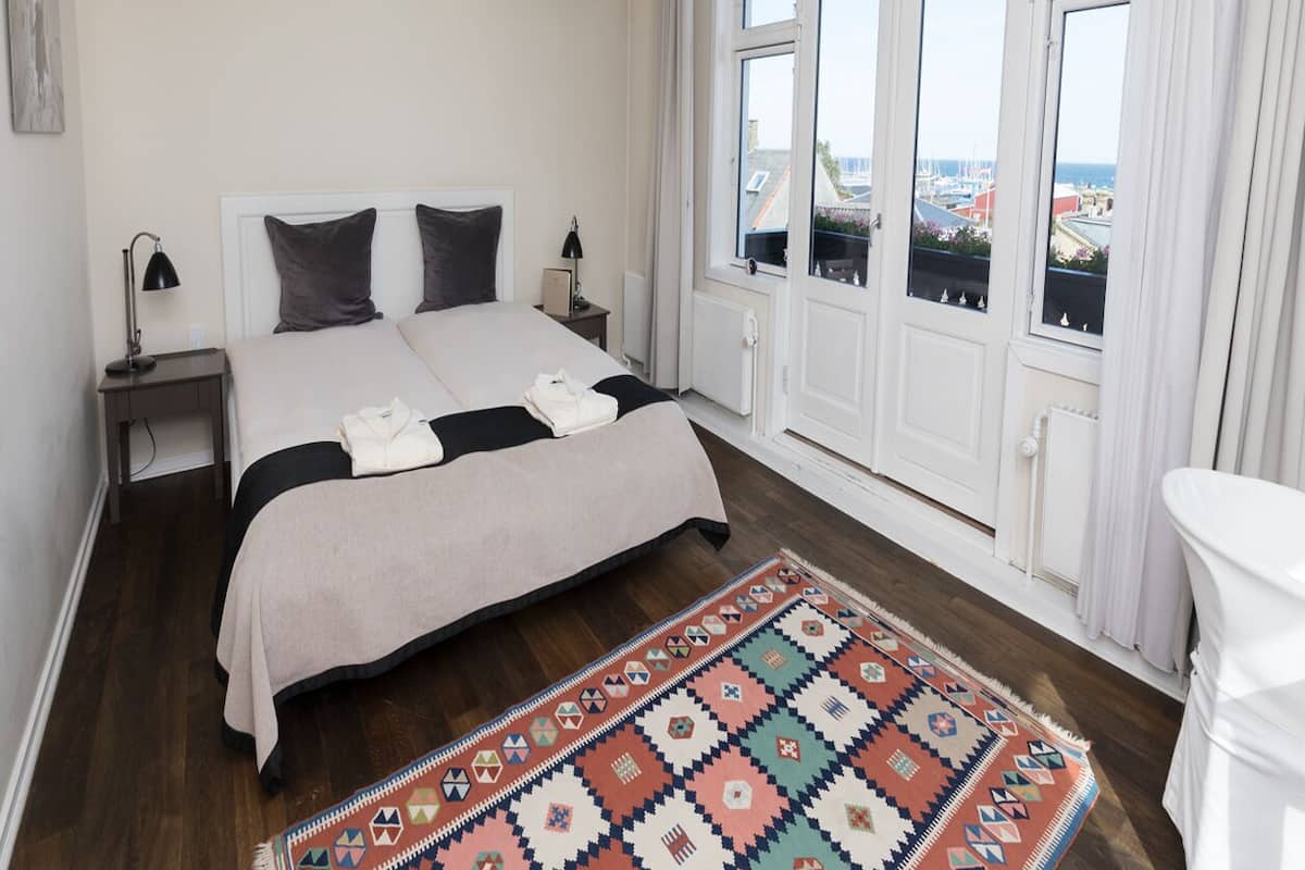 Un dormitorio con cama, mesita de noche, lámpara, ventana con vistas y alfombra estampada.