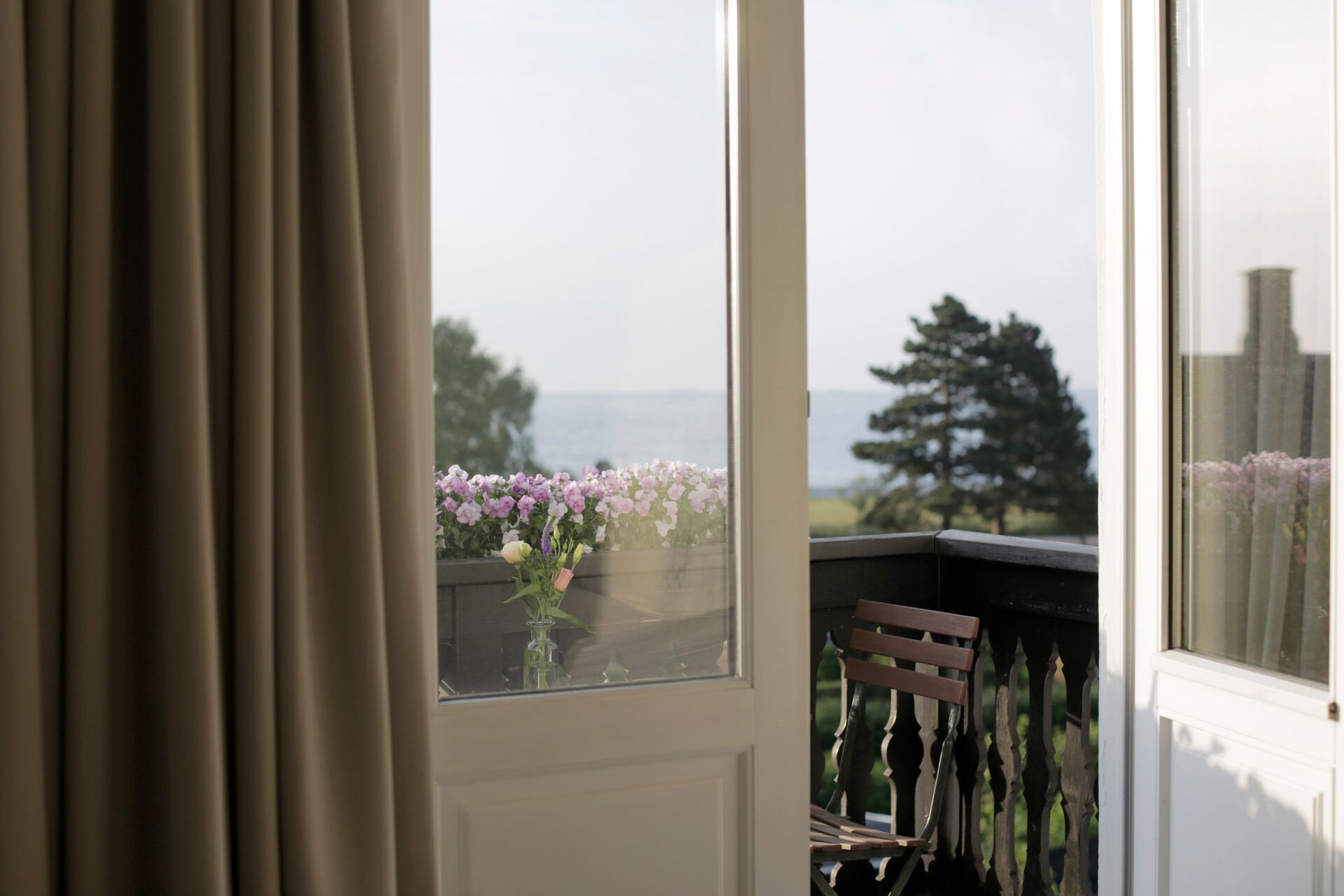 A balcony with a view of the sea, a chair, and a window with flowers.
