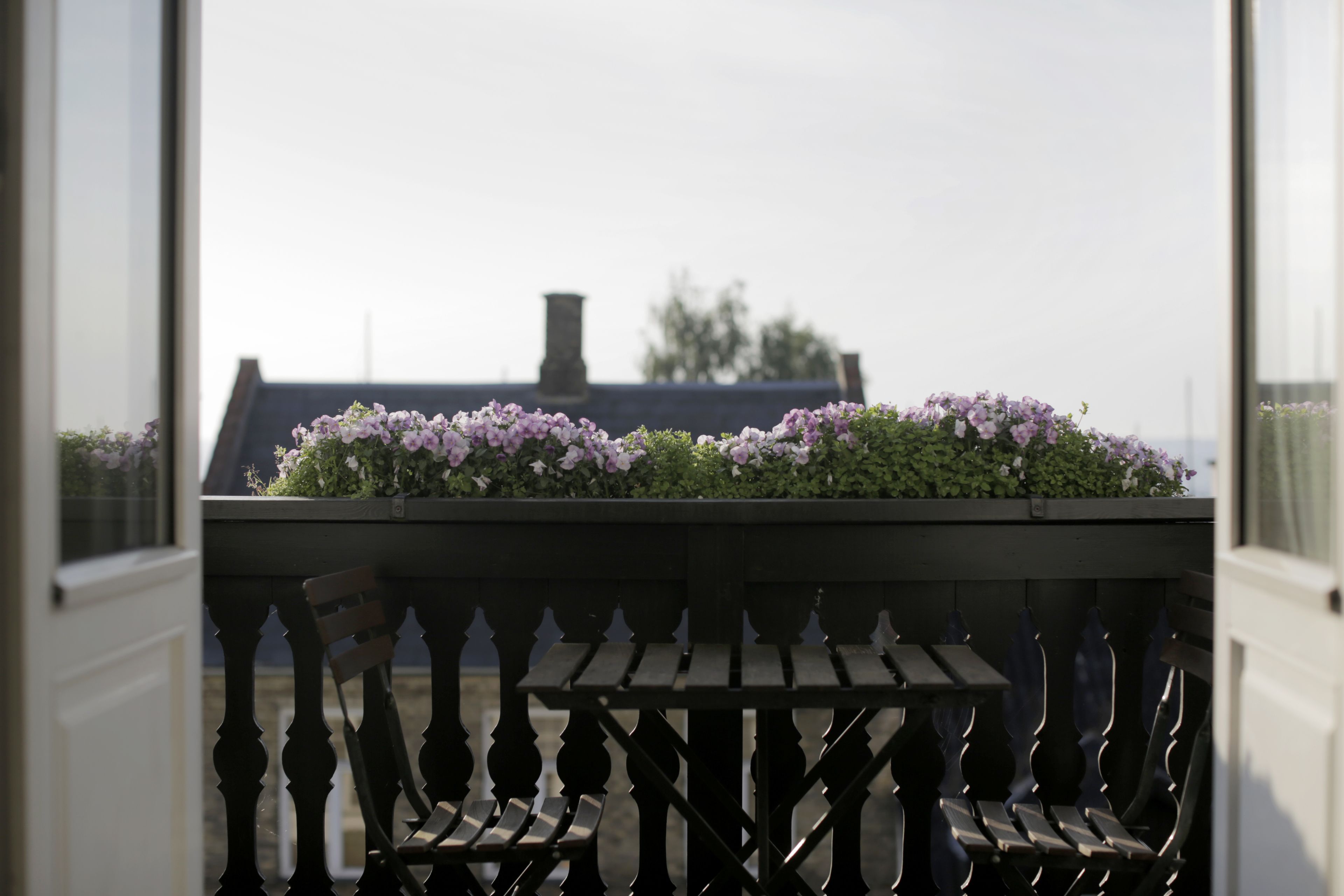 Un balcón con mesa y sillas, una jardinera con flores moradas, y vista a un edificio con chimenea.