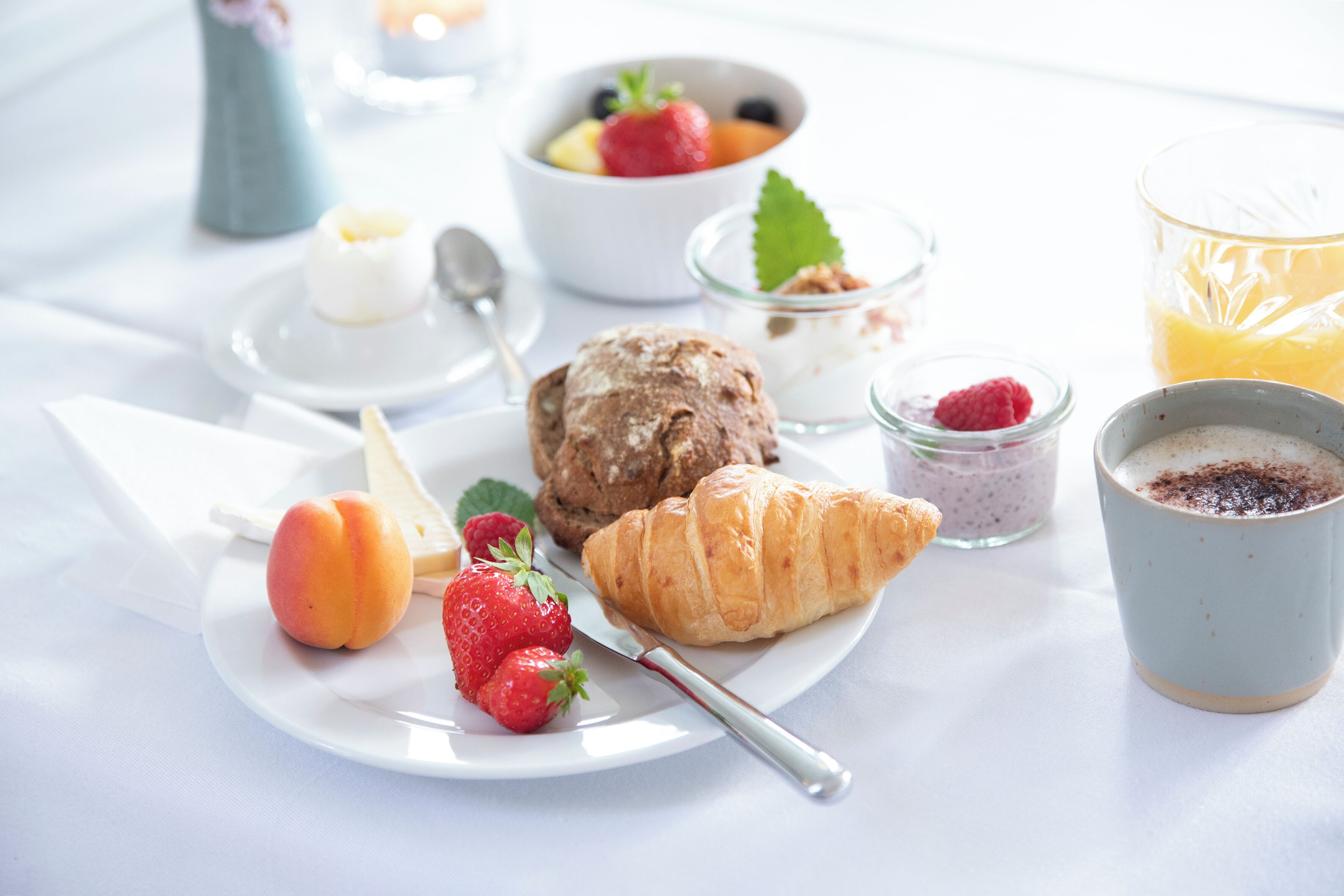 A breakfast setting with a croissant, strawberries, a peach, a bowl of fruit, a glass of orange juice, and a cup of coffee.