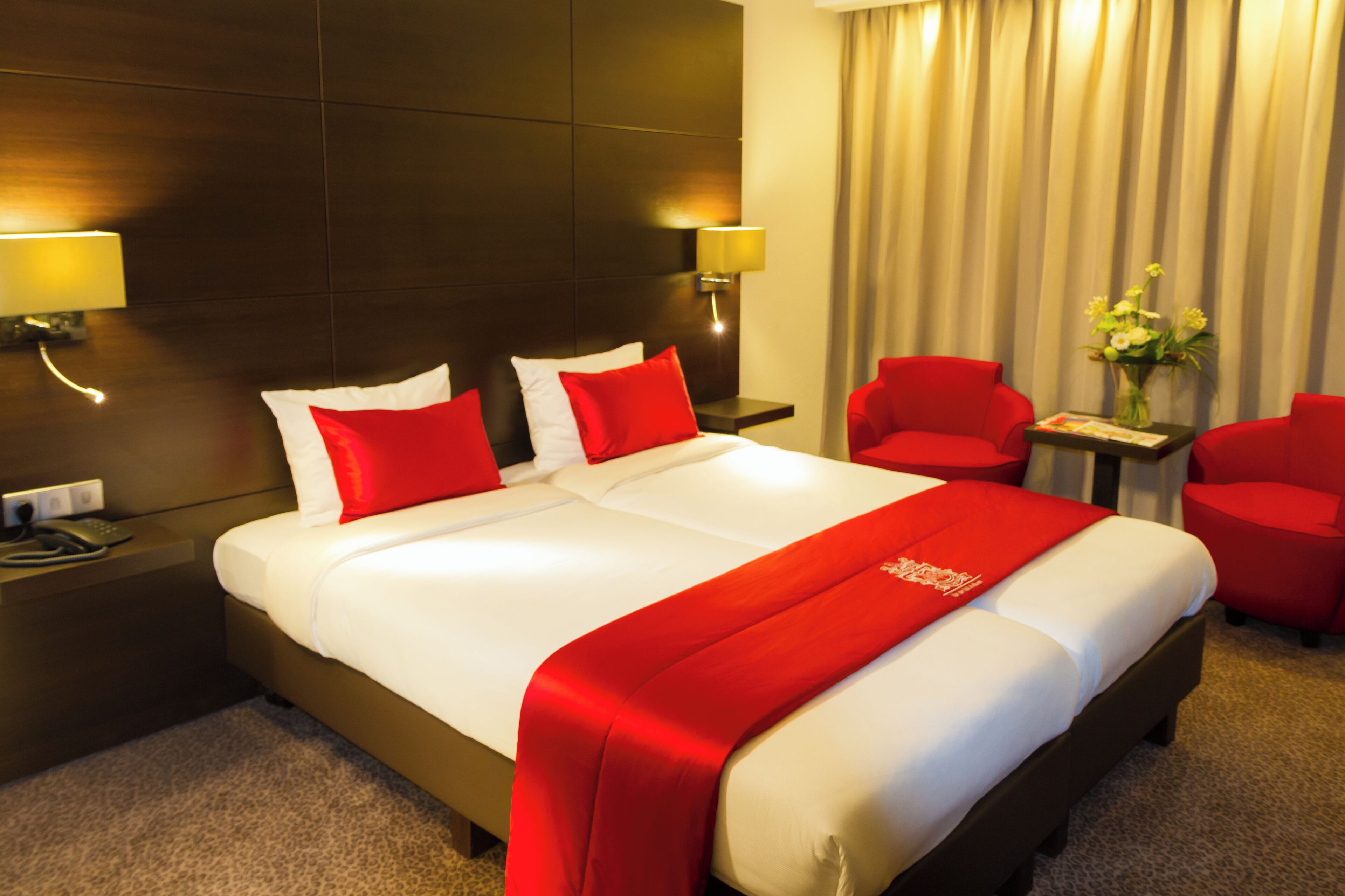 Une chambre d’hôtel avec un grand lit, deux fauteuils rouges, une petite table avec un vase et deux lampes fixées au mur.