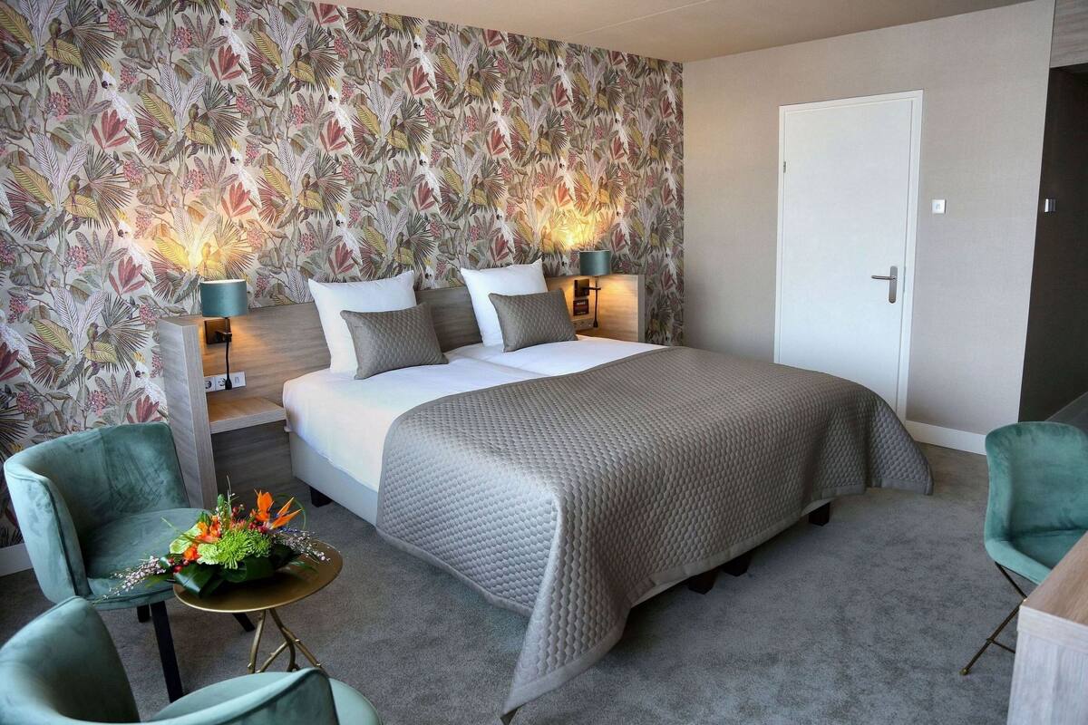 Une chambre d’hôtel avec un grand lit, deux tables de chevet avec des lampes, un fauteuil vert, une petite table ronde agrémentée d’un arrangement floral et un papier peint à motifs floraux.