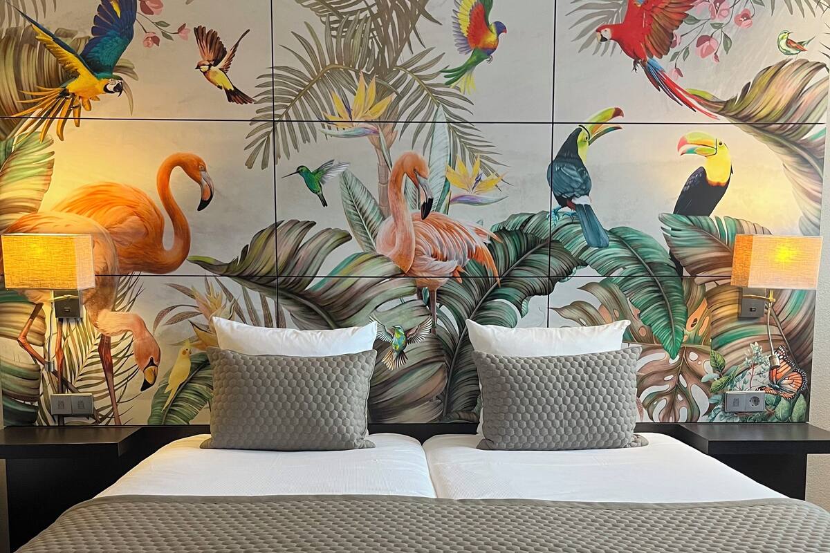 Une chambre d’hôtel comprenant un lit, deux oreillers, une table de chevet, une lampe et une fresque murale représentant des oiseaux et des plantes tropicales.