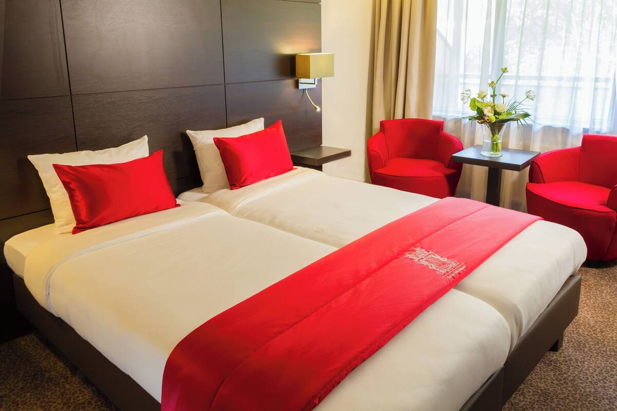 Une chambre d’hôtel avec deux lits, une literie rouge et blanche, une petite table sur laquelle est posé un vase de fleurs, et des chaises rouges.