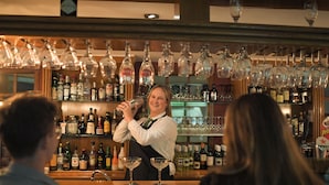 A bar area with a bartender preparing drinks, shelves of liquor bottles, and hanging wine glasses.