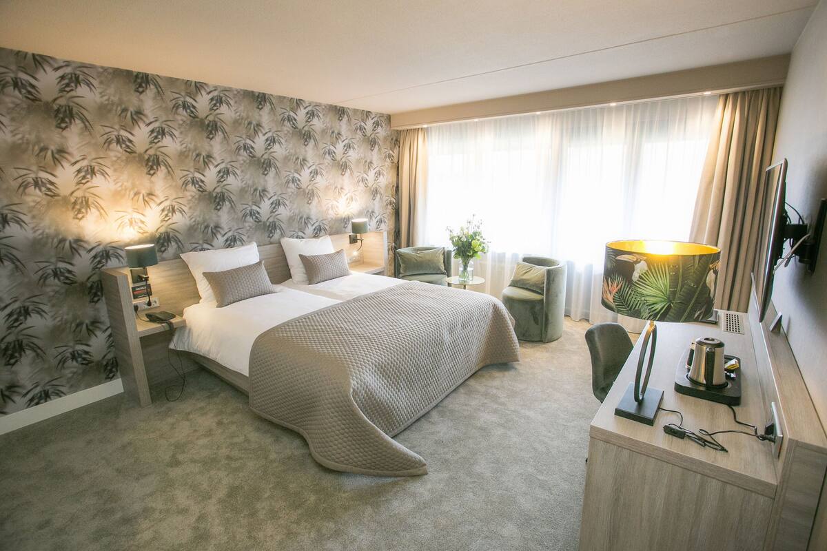 Une chambre d’hôtel avec un grand lit, un bureau avec une lampe, une chaise et une petite table sur laquelle est posée une plante.