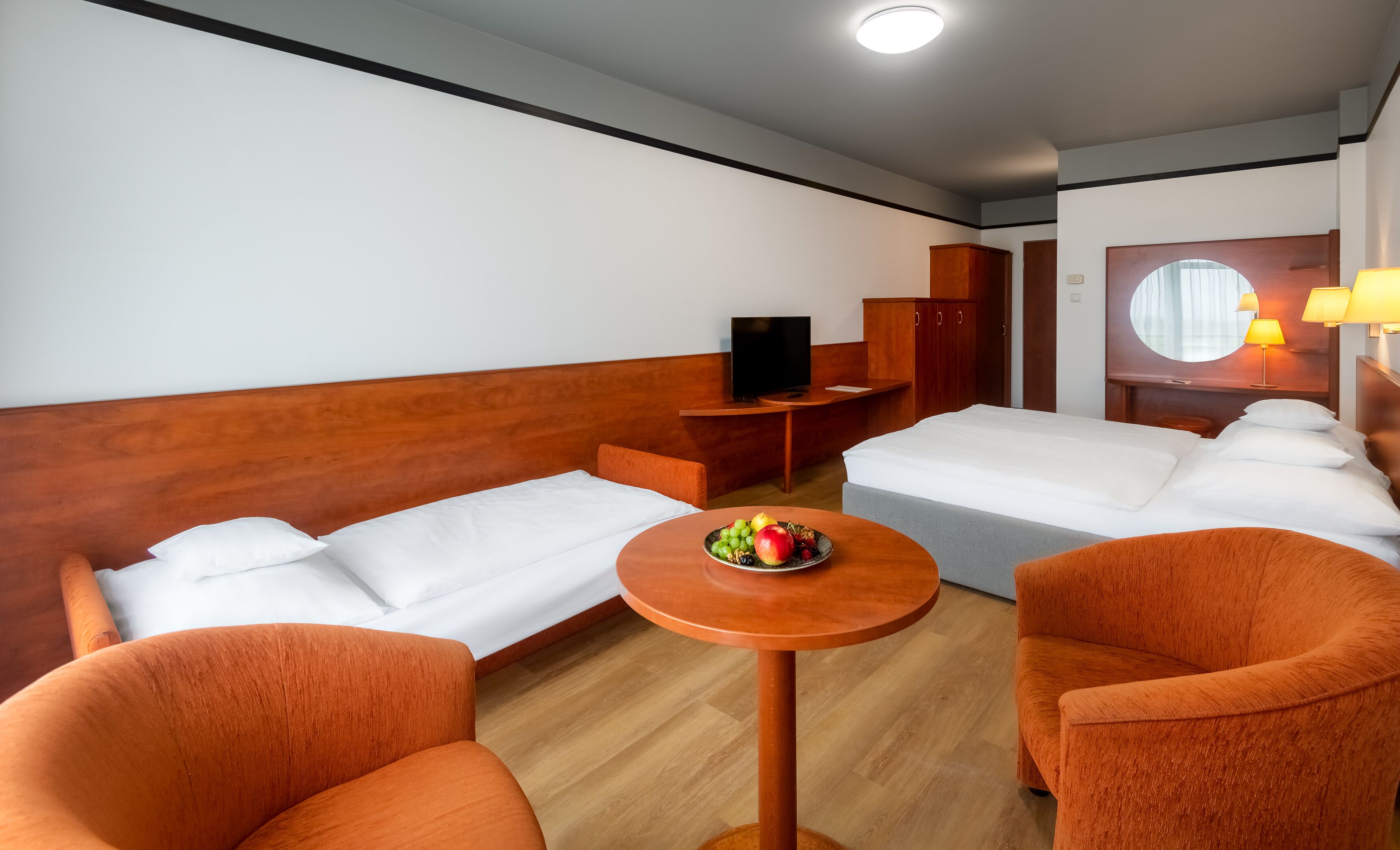 A hotel room with a bed, two orange armchairs, a small table with fruit, a TV, and a wardrobe.