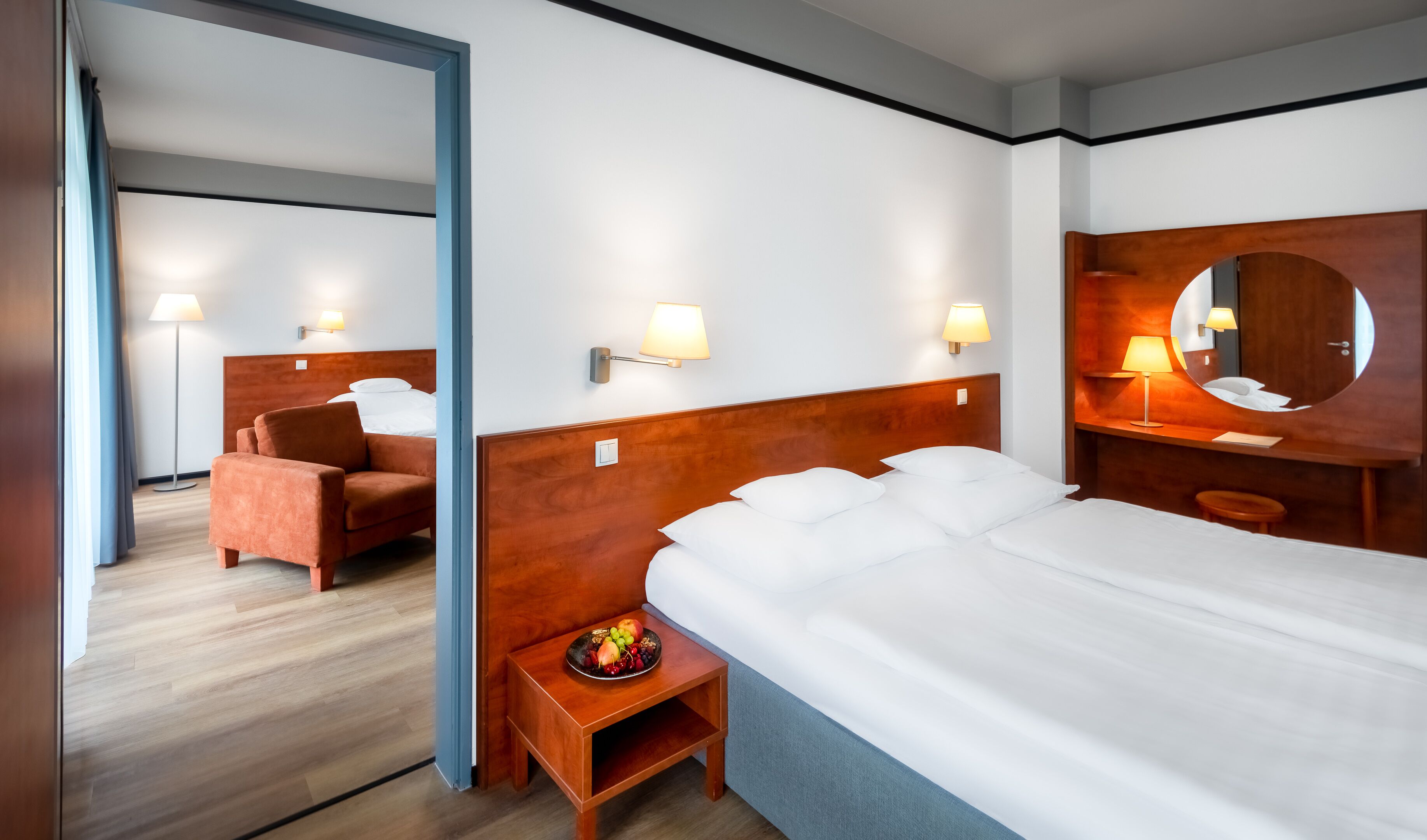 Quarto de hotel com uma cama grande, cabeceira de madeira, mesa de cabeceira com abajur, espelho e um espaço de estar com uma poltrona.