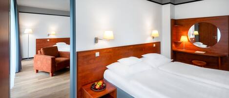 Una habitación de hotel con una cama grande, un cabecero de madera, una mesita de noche con lámpara, un espejo y una zona de estar con una silla.