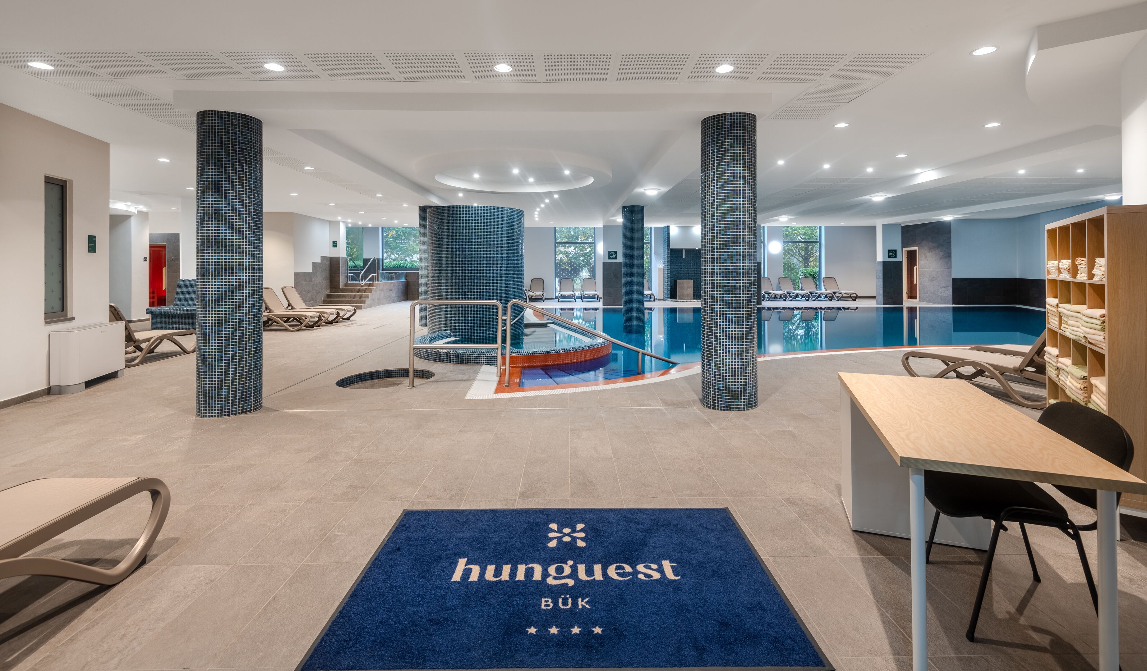 En modern hotellobby med ett poolområde, sittplatser och en receptionsdisk.