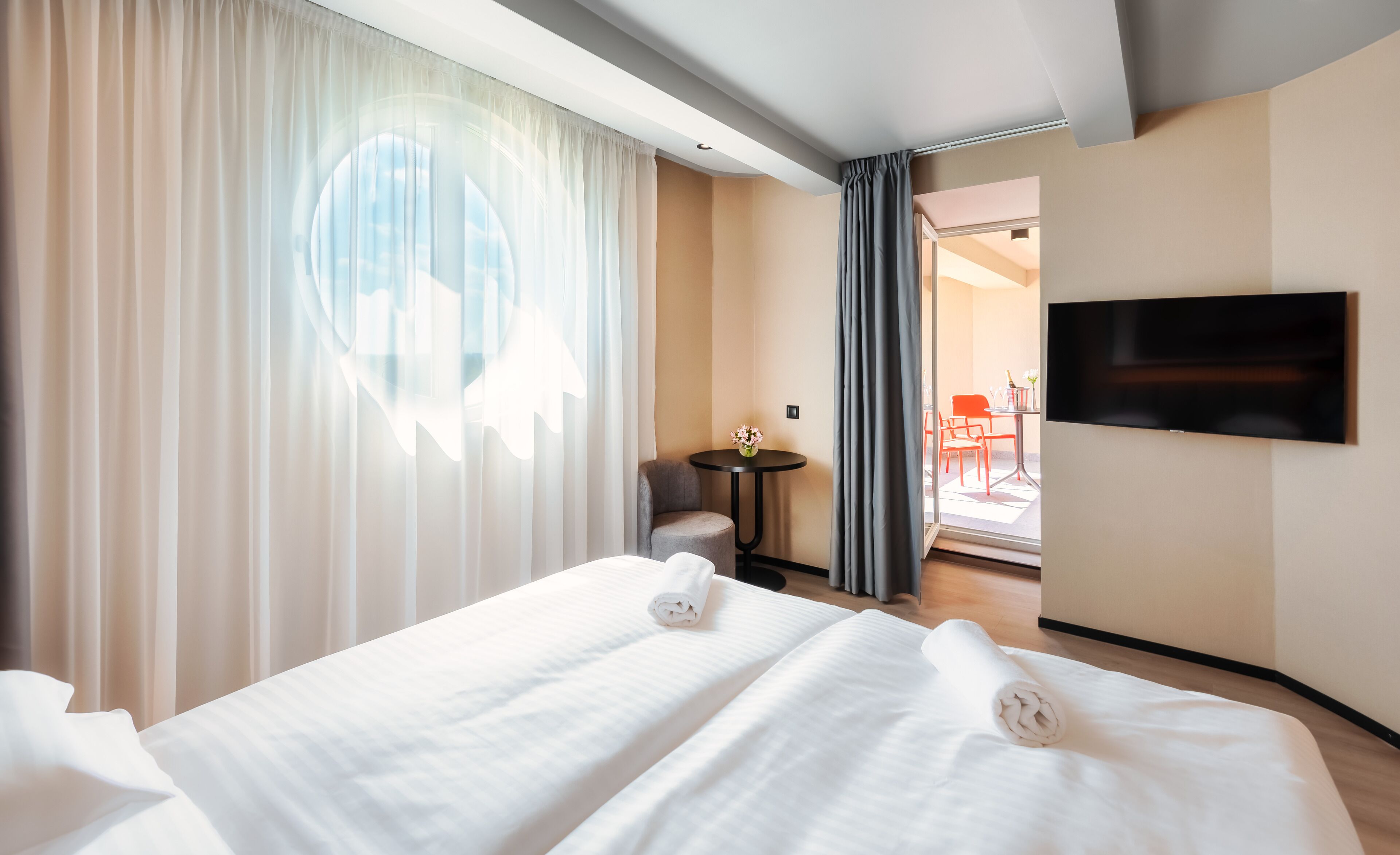 Una habitación de hotel con cama, una mesita con un florero, una silla y un televisor montado en la pared.