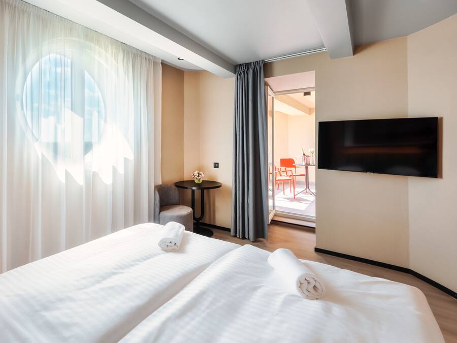 Una habitación de hotel con cama, una mesita con un florero, una silla y un televisor montado en la pared.