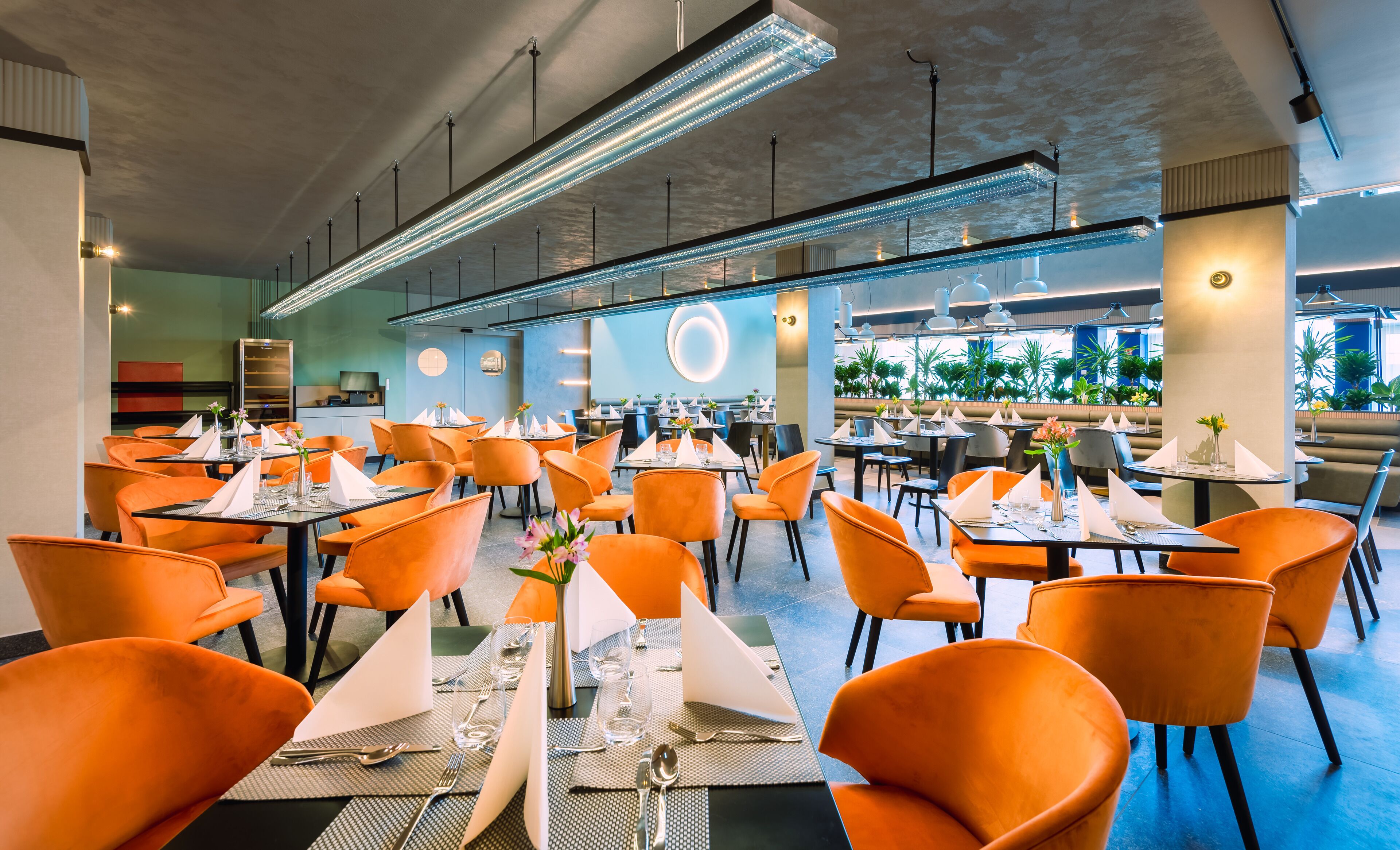 A spacious restaurant with orange chairs and tables, modern lighting, and a view of greenery outside.