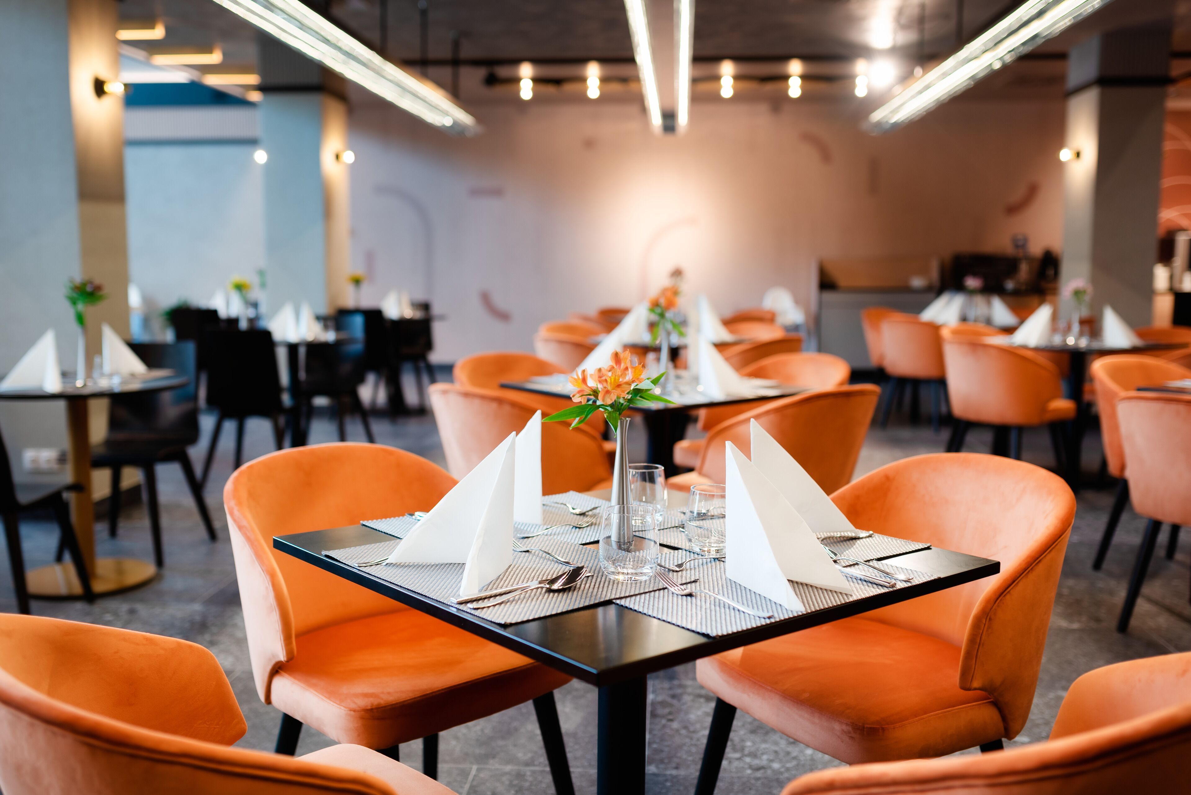 A restaurant with orange chairs and black tables, each set with a white napkin, a glass, and cutlery.
