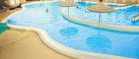 A large, curved swimming pool with two thatched umbrellas, surrounded by trees and a sandy area.