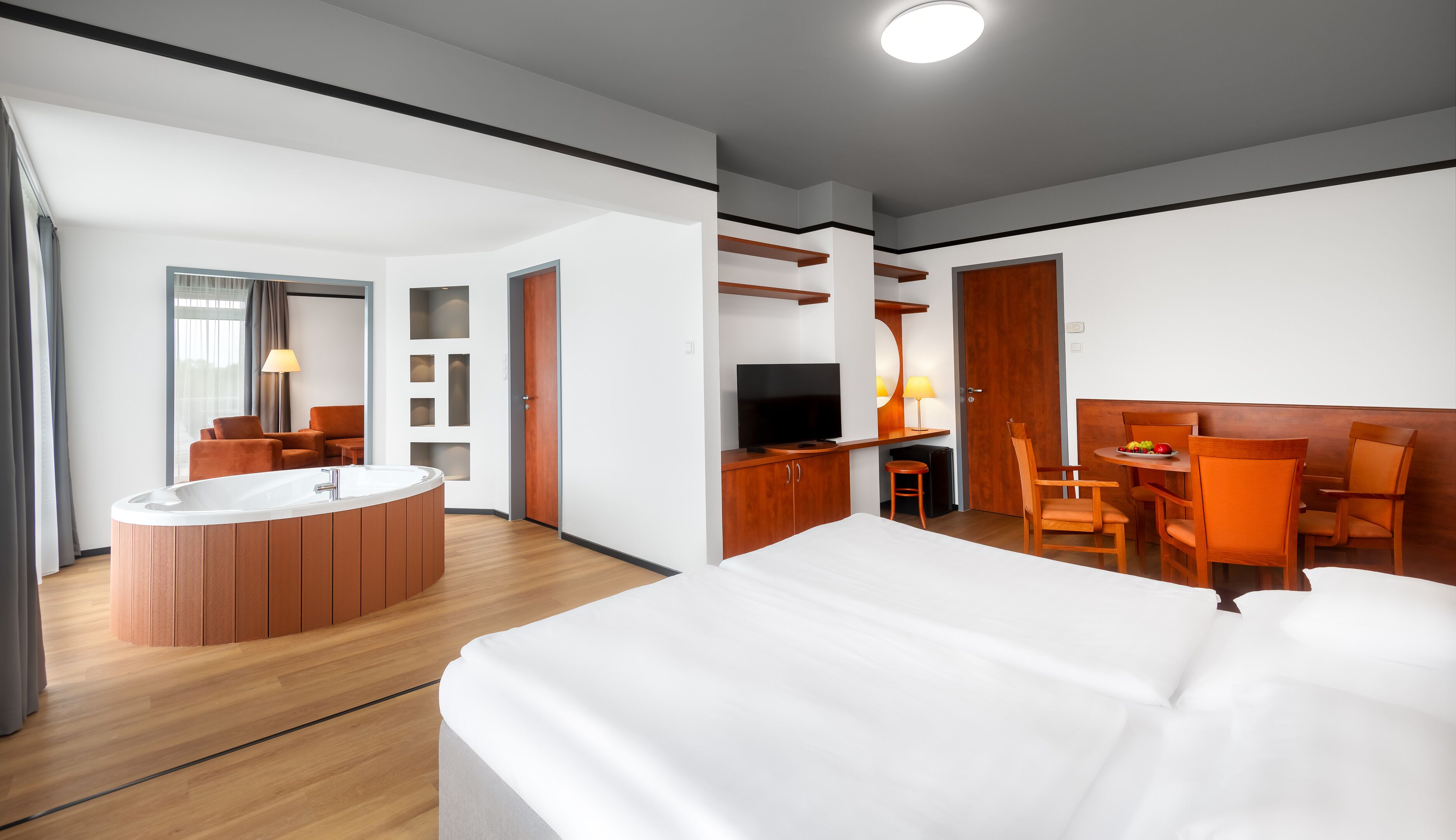 Quarto de hotel moderno com uma cama grande, área de estar separada com mesa e cadeiras, televisão e uma pequena cozinha.