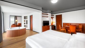 Quarto de hotel moderno com uma cama grande, área de estar separada com mesa e cadeiras, televisão e uma pequena cozinha.