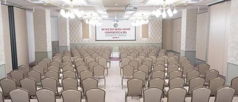 Une salle de conférence avec des rangées de chaises, un écran de projection et une banderole pour l’hôtel Hunguest Fenyő, qui propose des services de conférence et de spa.