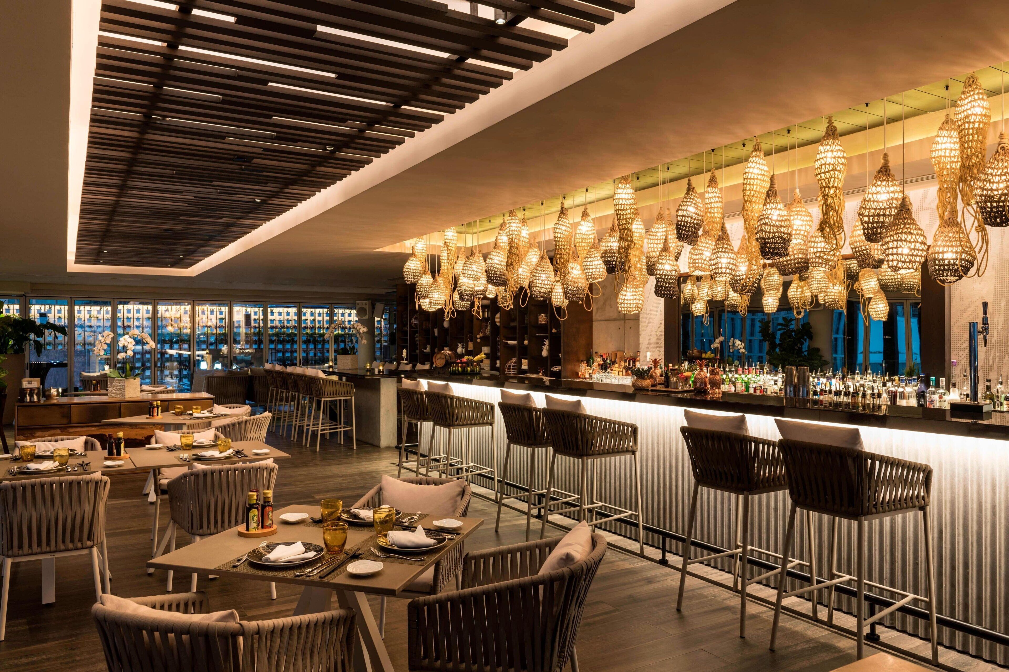 A modern bar area with high stools, a long counter, and hanging decorative lights.