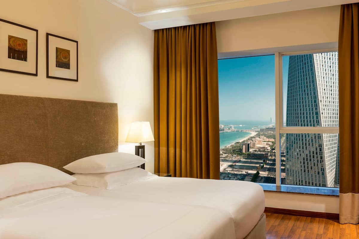 A hotel room with a large bed, a nightstand, a lamp, and a view of a cityscape through a window.