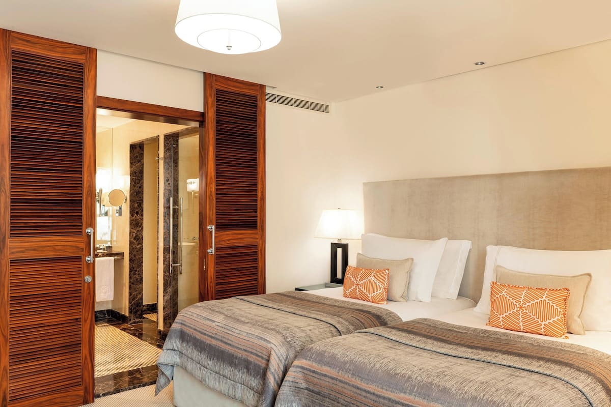 A hotel room with a large bed, a nightstand with a lamp, a wooden wardrobe, and a bathroom visible through an open door.