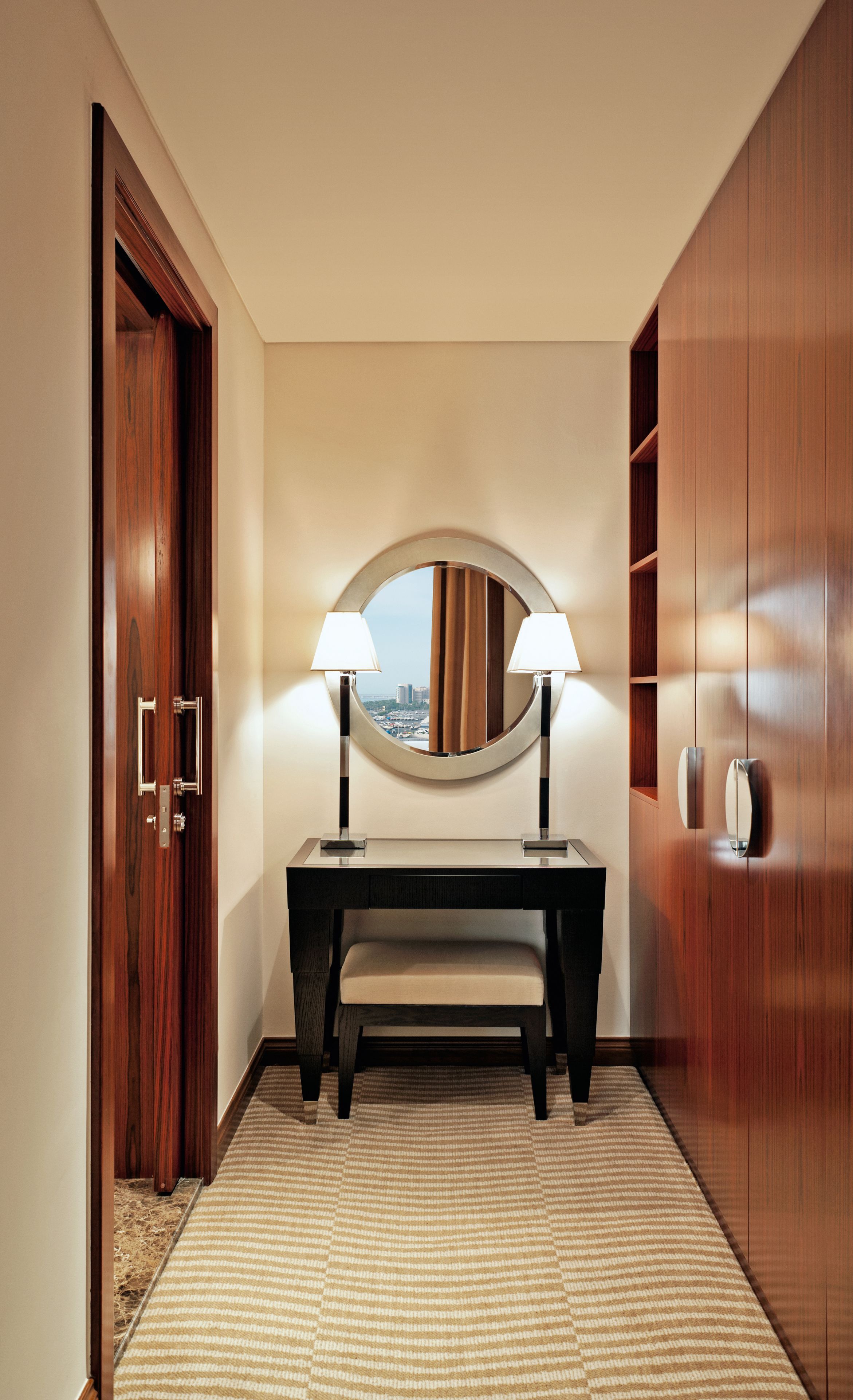 Un pasillo con una puerta de madera, un tocador con espejo, dos lámparas y un banco.