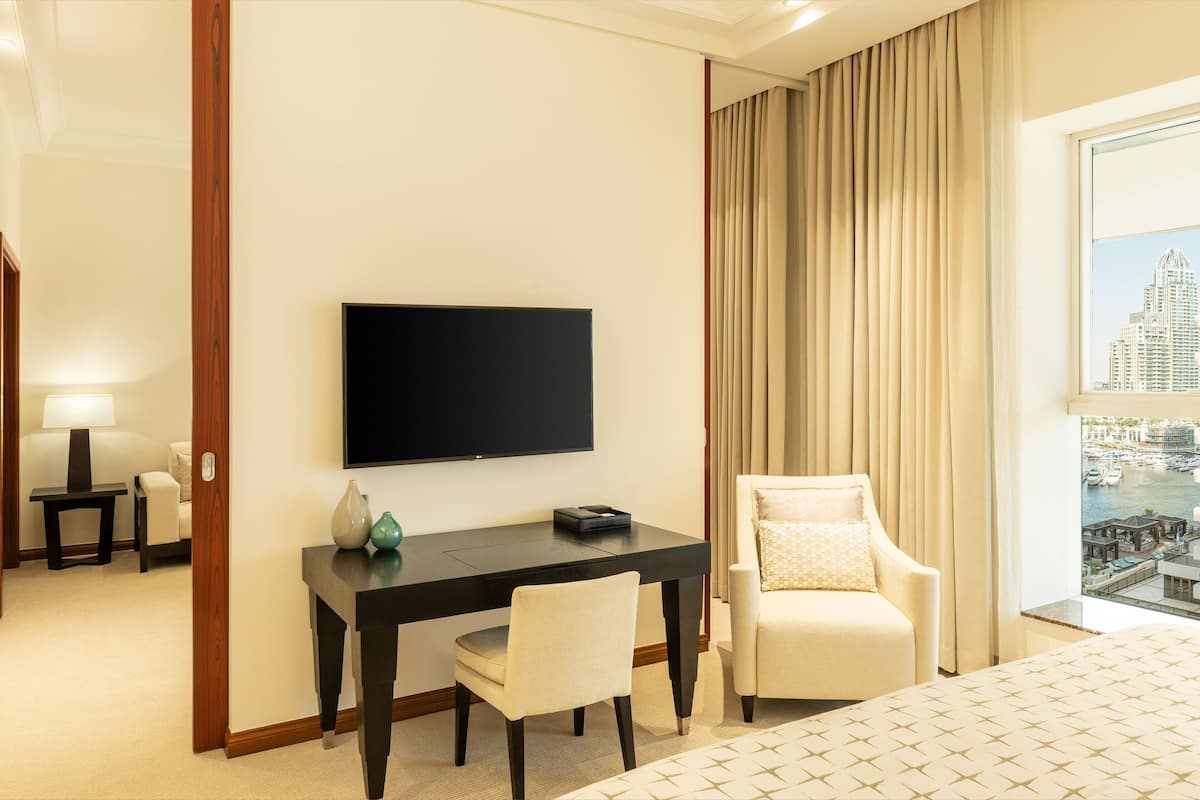 A hotel room with a bed, a desk with a chair, a flat-screen TV, a lamp, and a view of a cityscape through a large window.