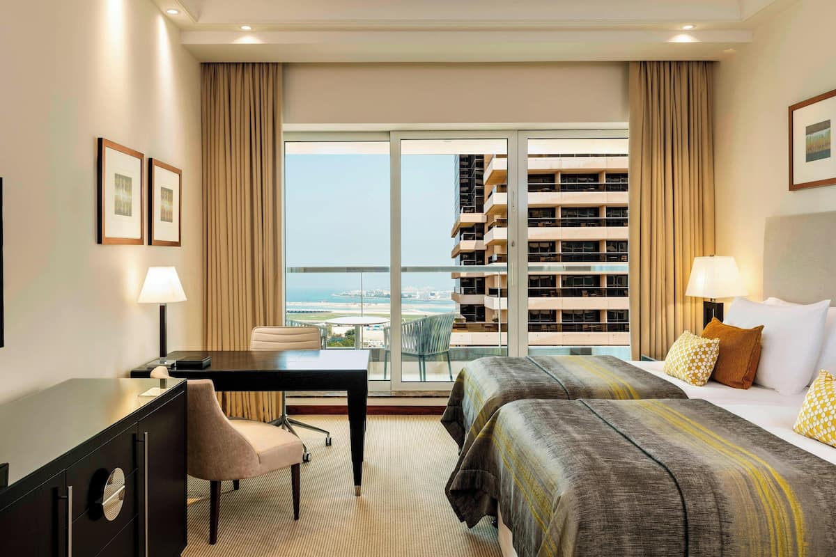 A hotel room with a desk, two beds, a chair, and a large window with a view of a high-rise building and the sea.