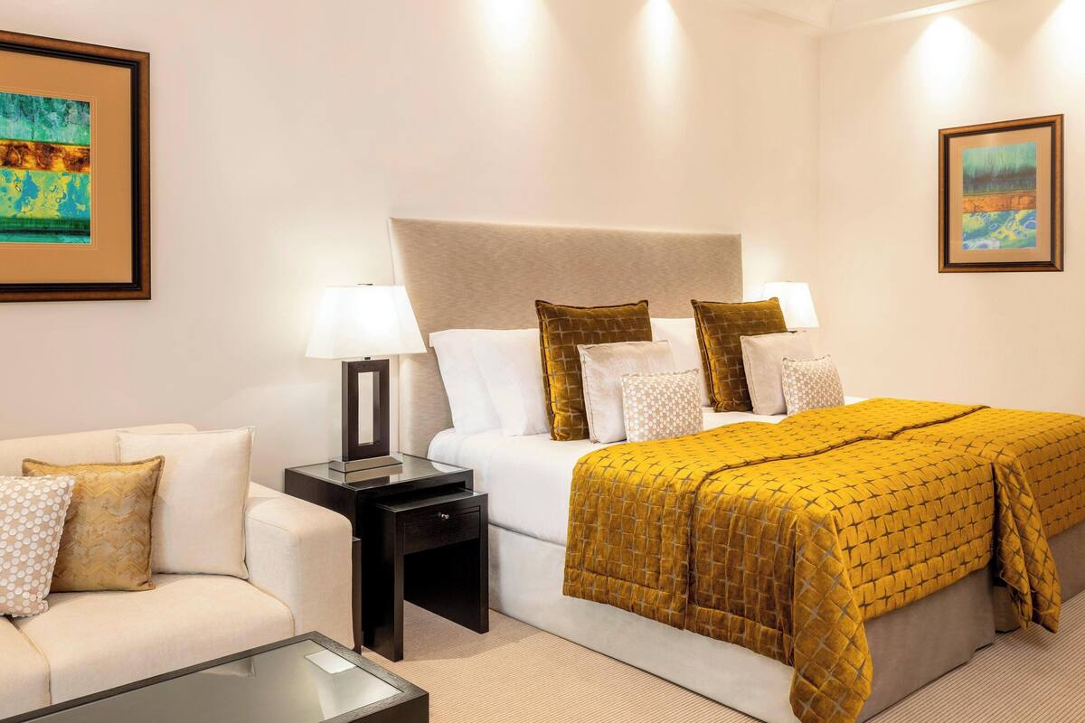 A hotel room with a bed, a nightstand, a bedside table, a sofa, and two framed artworks on the walls.