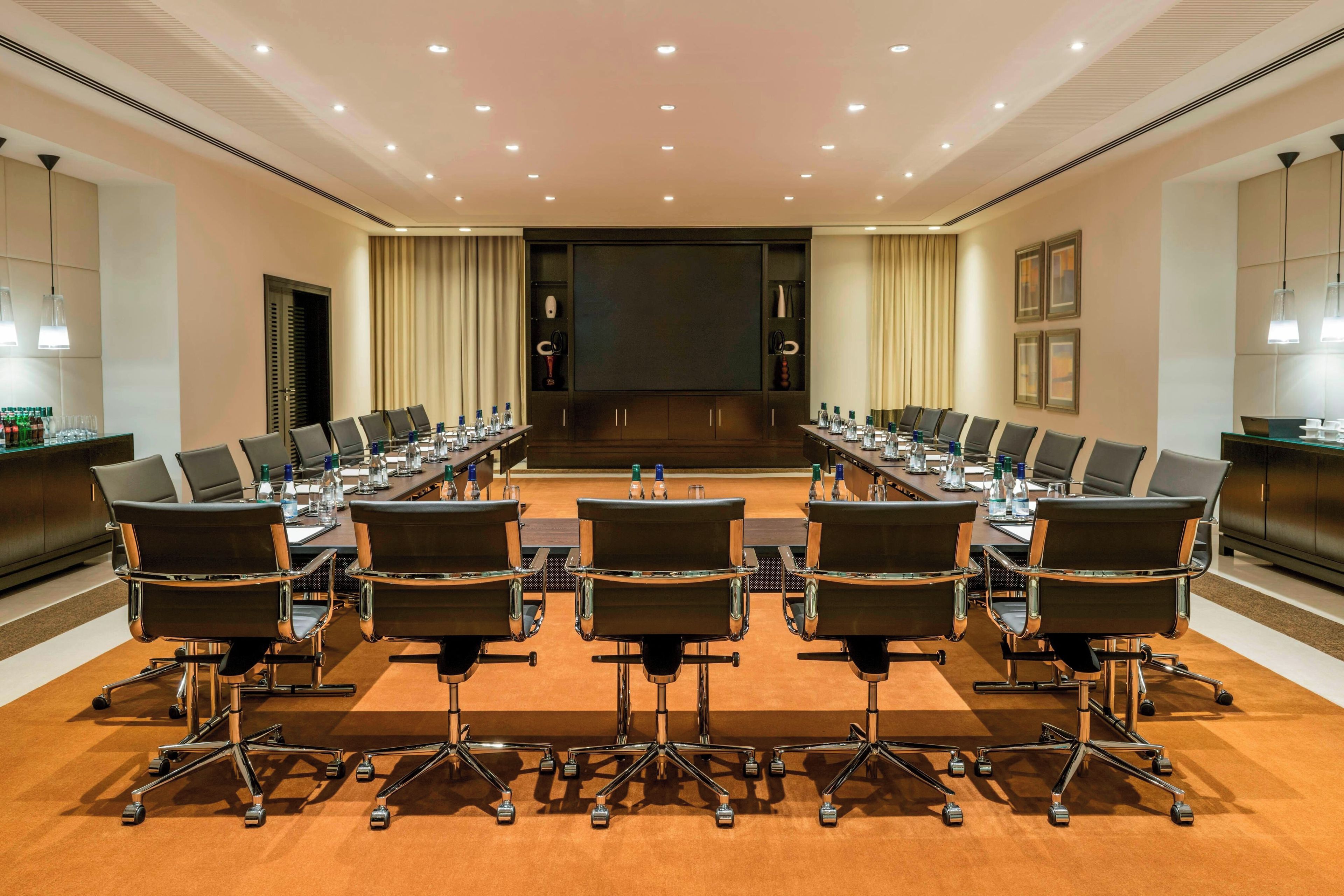 Un salón de conferencias con una larga mesa, varias sillas y una pantalla grande.