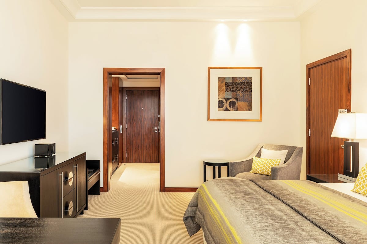 A hotel room with a bed, a TV, a desk, and a chair. There is a door leading to another room and a framed picture on the wall.