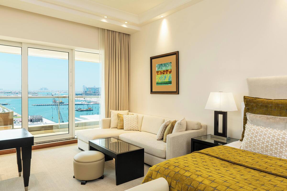 A hotel room with a large window offering a city view, a sofa, a bed, a coffee table, and a lamp.