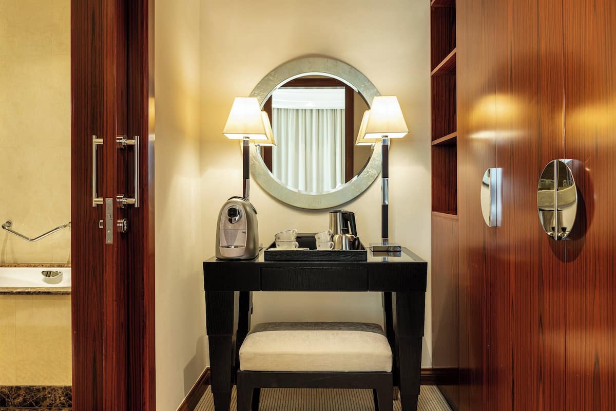 A hotel room with a wooden vanity, a round mirror, two wall-mounted lamps, a coffee maker, and a small bench.