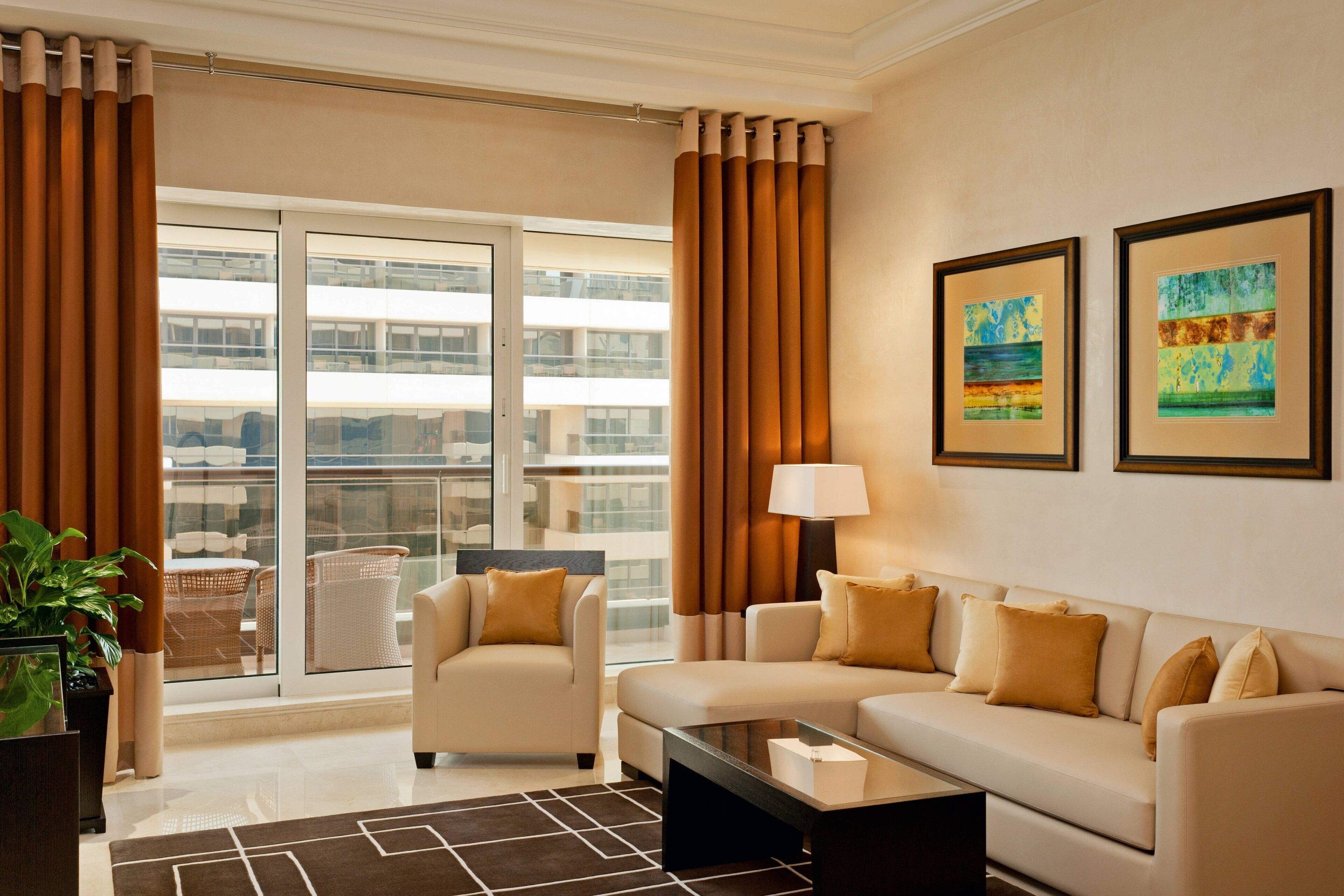 A modern living room with a sofa, armchair, and coffee table. There is a large window with curtains, a view of buildings outside, and two framed artworks on the wall.
